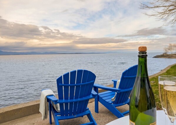 A table with a bottle of wine and glasses, two blue chairs, and a view of a body of water with mountains in the distance.