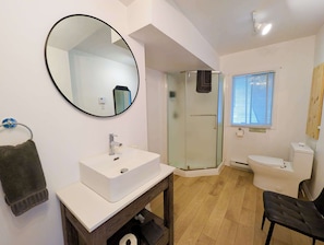 A bathroom with a white sink, a round mirror, a glass shower enclosure, a toilet, and a window with blinds.