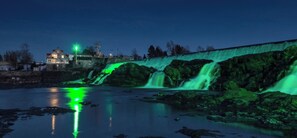 Une cascade illuminée de lumiÚre verte, un mur de pierre et un bùtiment en arriÚre-plan.
