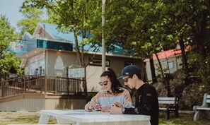 Deux personnes sont assises Ă une table en plein air, avec un bĂątiment visible en arriĂšre-plan.