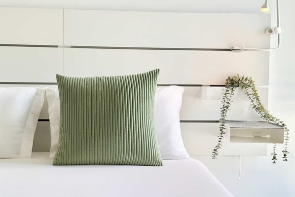 Una cama con un almohadón verde y ropa de cama blanca, un cabecero blanco y una planta pequeña al lado.