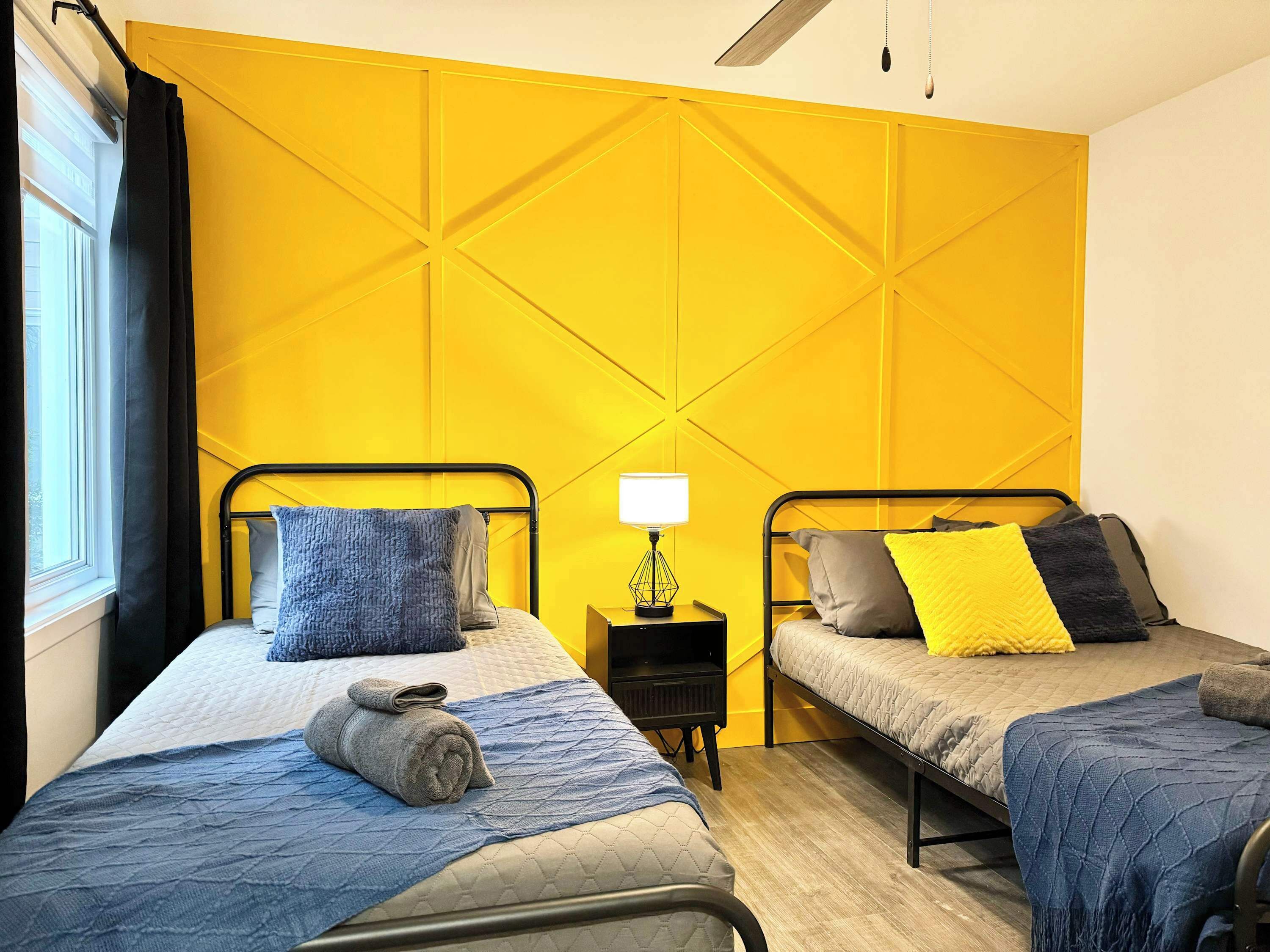 A room with two beds, a nightstand with a lamp, and a window with curtains.