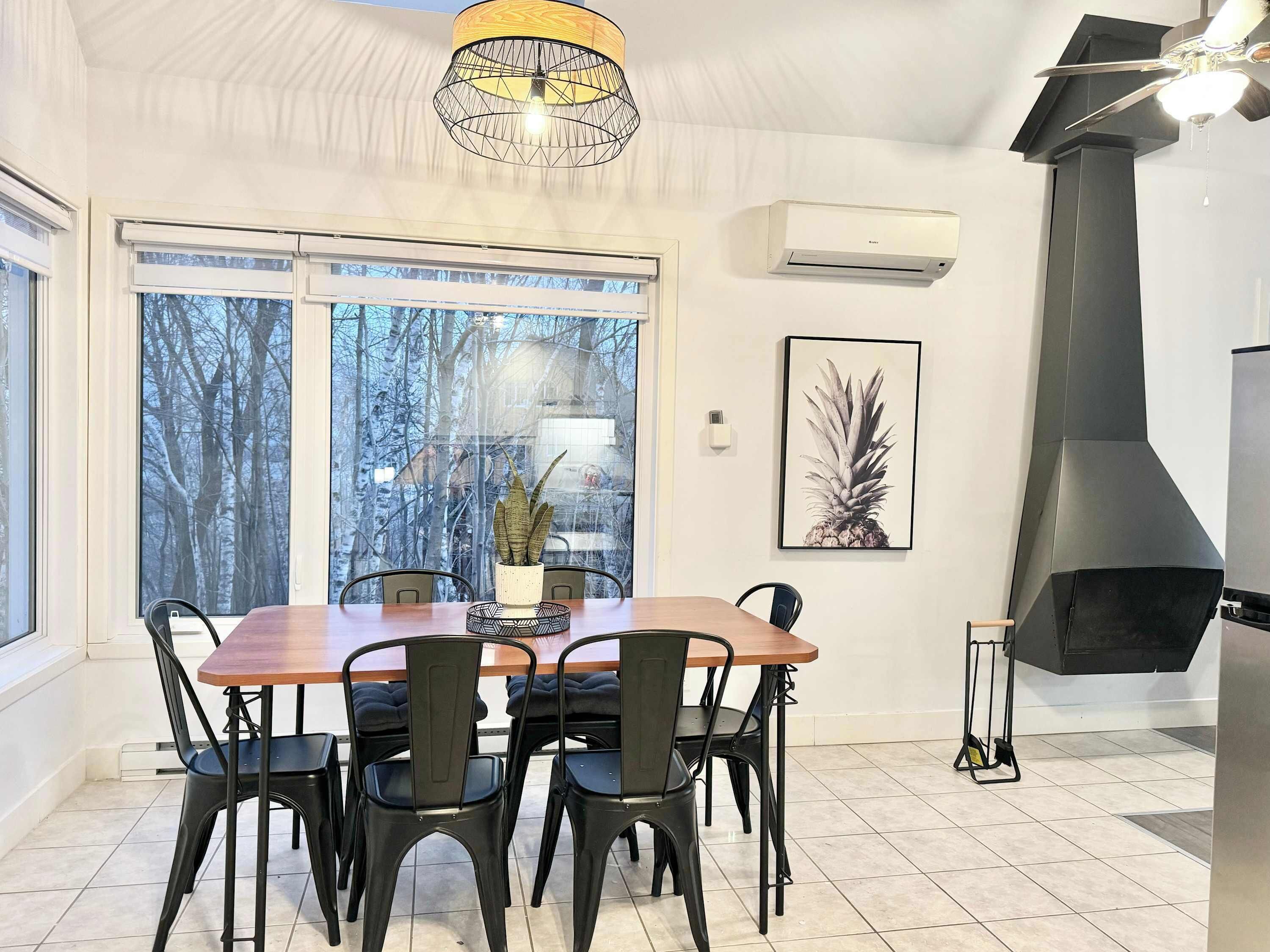 A dining area with a wooden table and black chairs, a large window with a view of trees, a wall-mounted air conditioner, and a black stove.