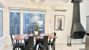 A dining area with a wooden table and black chairs, a large window with a view of trees, a wall-mounted air conditioner, and a black stove.