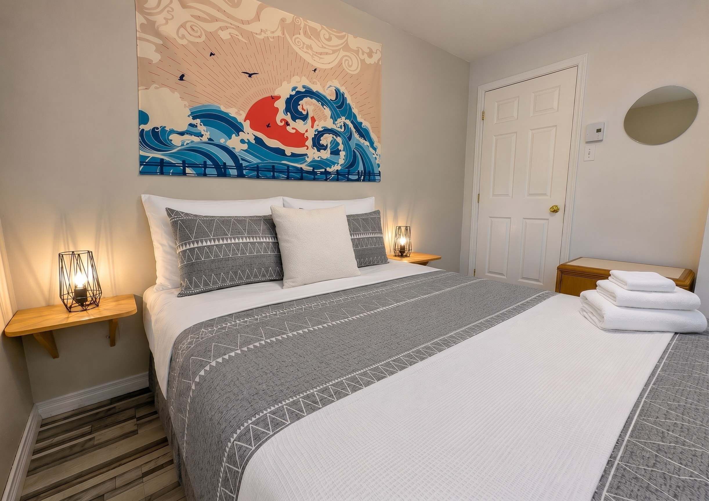 A hotel room with a bed, nightstands, a wall art of waves, a door, and a mirror.