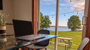 A room with a glass table, a laptop, a chair, a potted plant, and a view of a lake with trees and a picnic table.