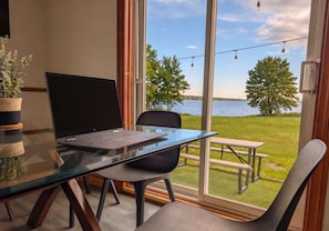 A room with a glass table, a laptop, a chair, a potted plant, and a view of a lake with trees and a picnic table.