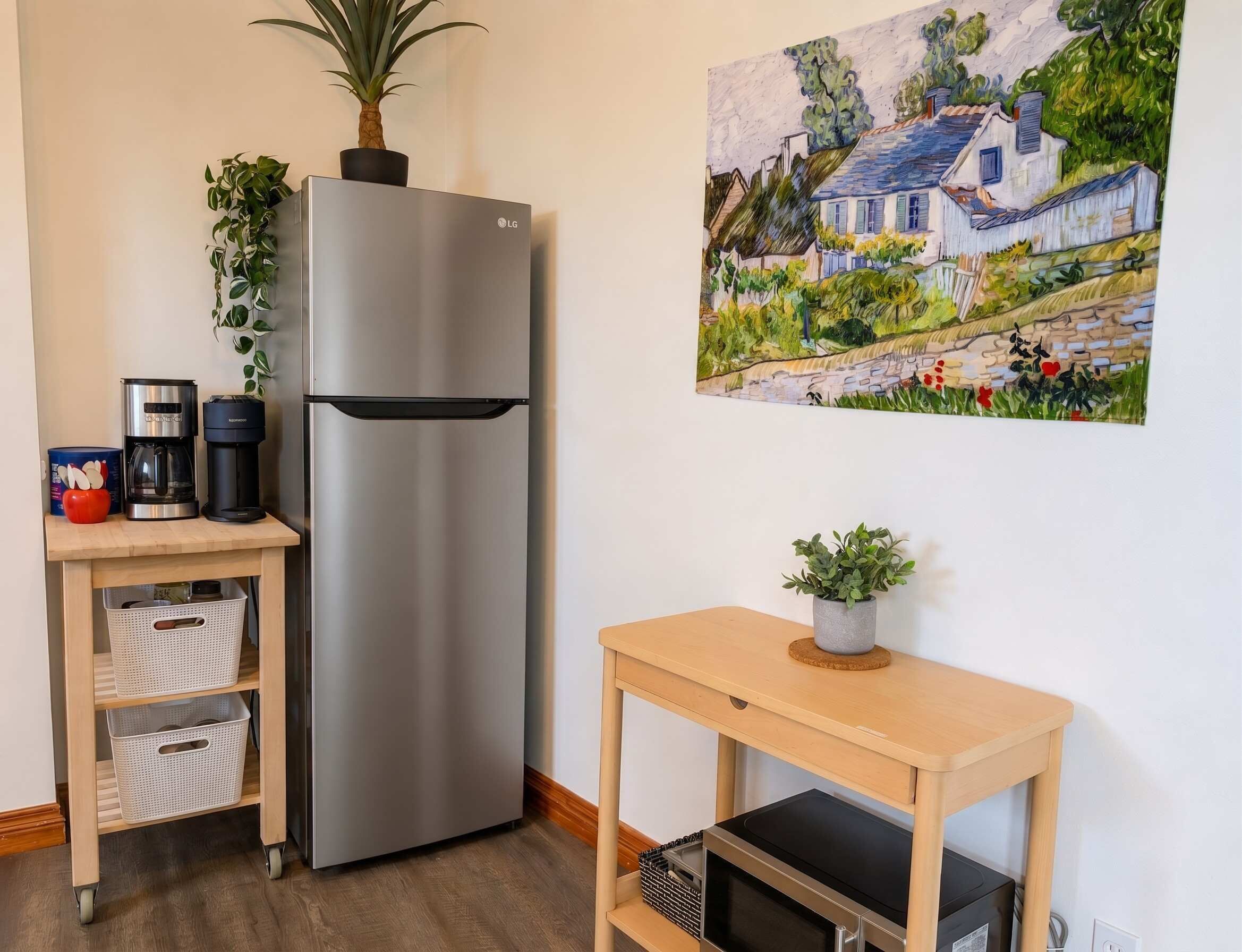 A kitchen with a stainless steel refrigerator, a coffee maker, a microwave, a small table with a plant, and a painting of a house on the wall.
