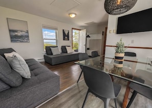 A modern living room with a glass dining table, grey sofas, and a view of the outdoors.
