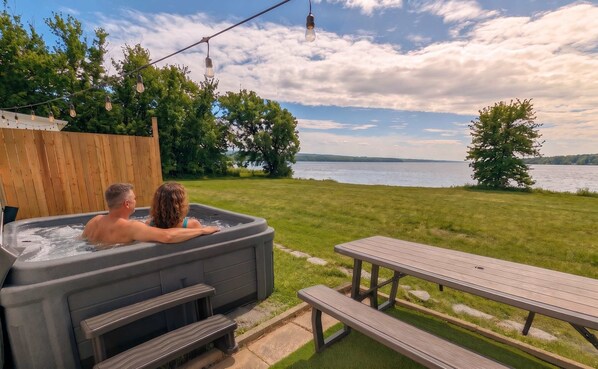 A hot tub with two people, a wooden deck, a picnic table, and a lake view.
