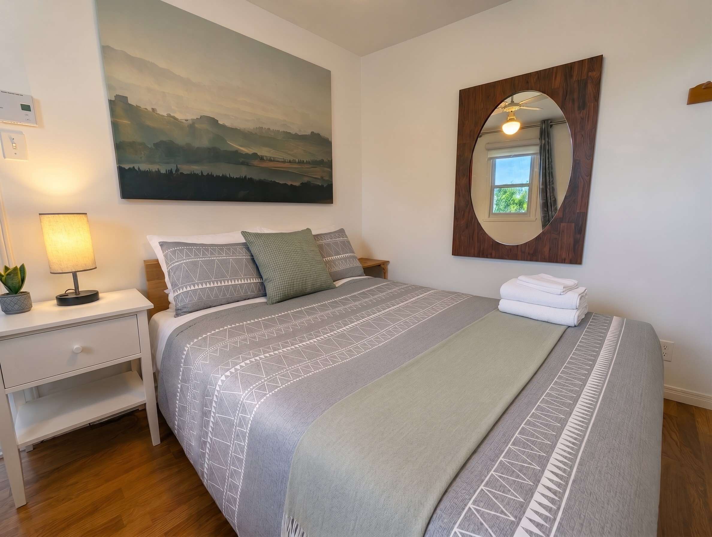 Ein ordentlich eingerichtetes Schlafzimmer mit Bett, Nachttisch, Lampe und einem großen Landschaftsbild.