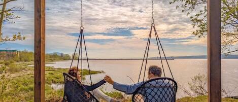 Dos personas en una silla colgante con vista a un cuerpo de agua y un puente distante.