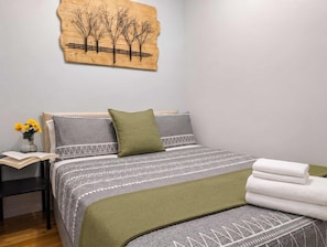 Ein ordentlich eingerichtetes Schlafzimmer mit einem Bett, Kissen, einem Nachttisch mit Buch und Blumen sowie einem Wandbild.