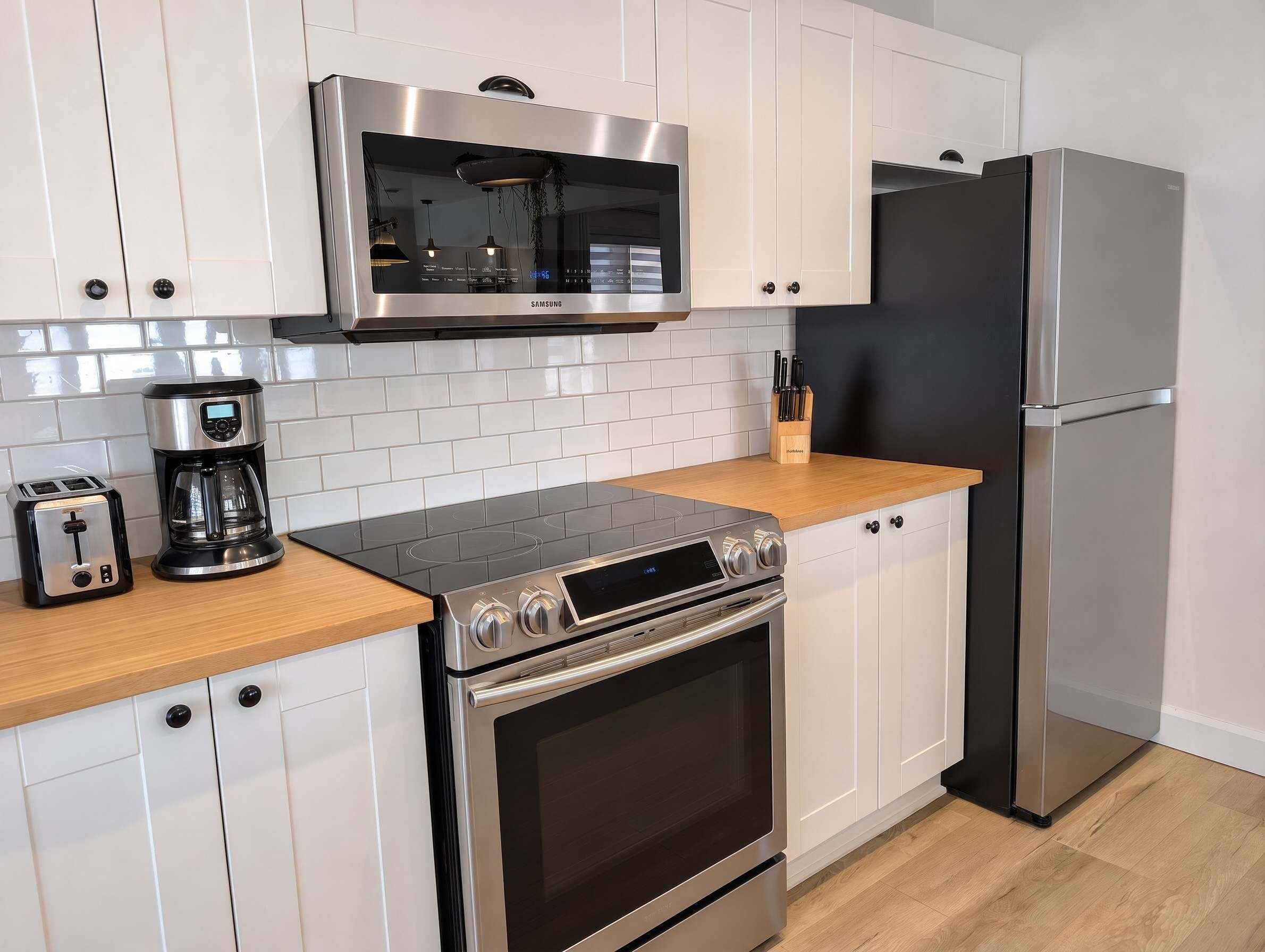 A modern kitchen with white cabinets, a stainless steel microwave, a coffee maker, a toaster, an oven, and a refrigerator.
