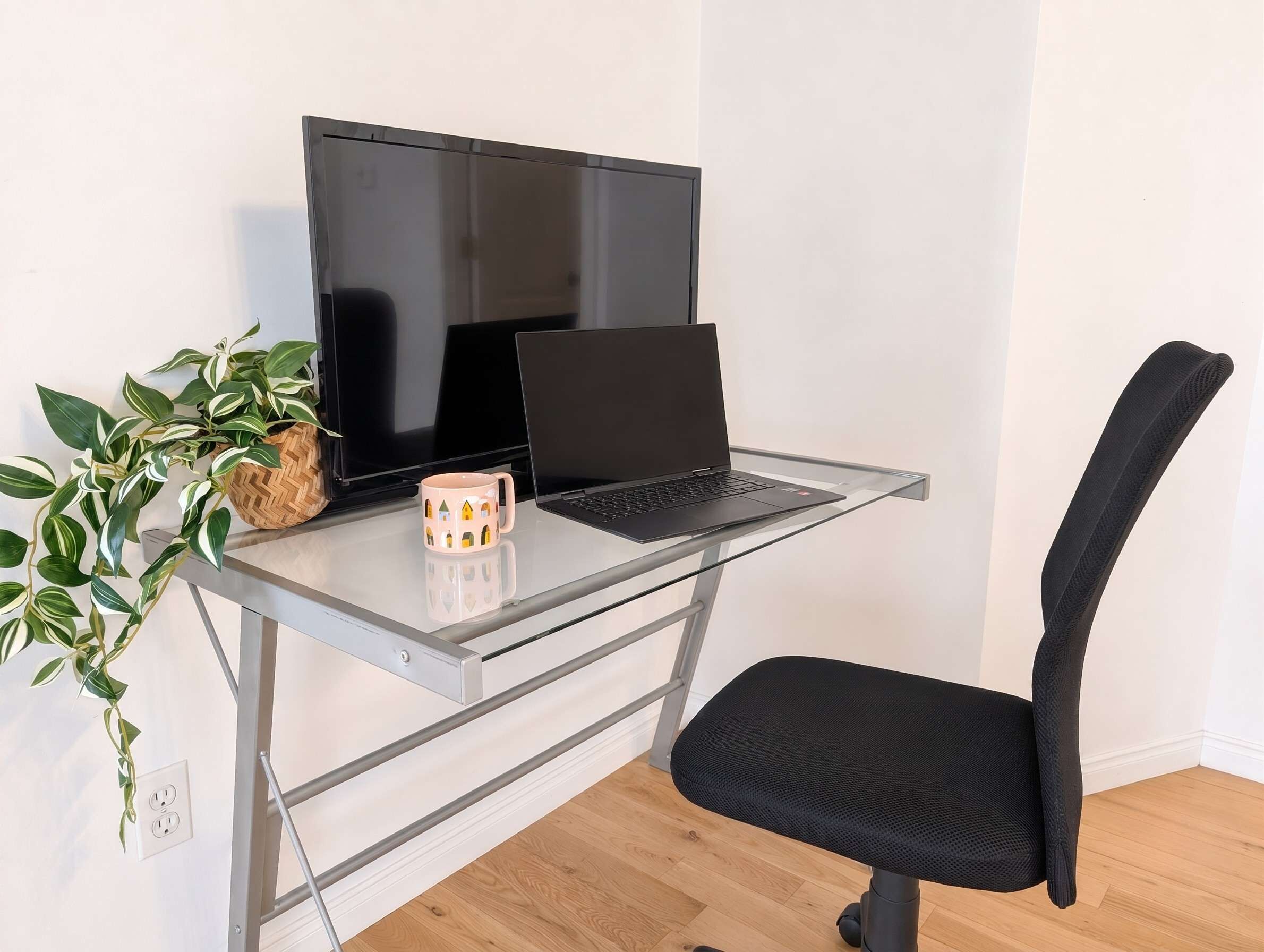 A workspace with a laptop on a glass desk, a black chair, and a potted plant.