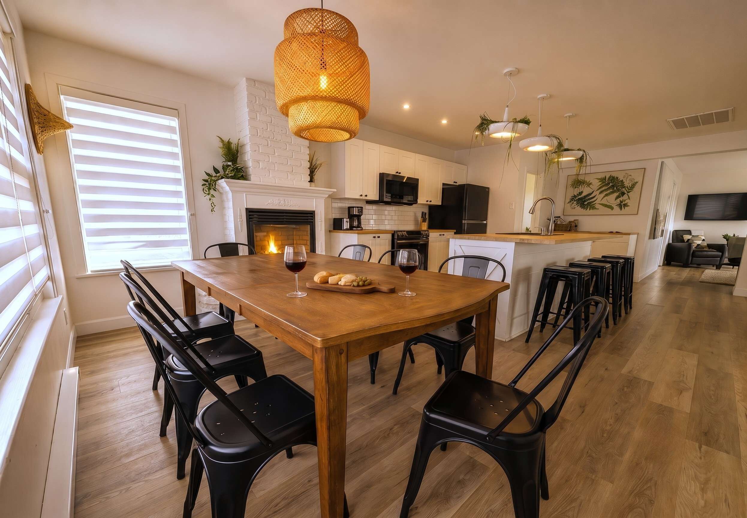 A dining area with a wooden table, black chairs, a hanging light fixture, and a kitchen in the background.