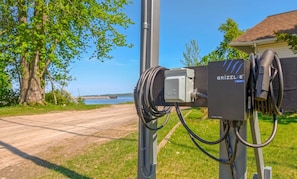 Une borne de recharge électrique portable pour véhicules, dotée d’un câble et d’un boîtier portant l’inscription « GRIZZLE DUO », installée au bord d’une route offrant une vue sur un plan d’eau et des arbres.