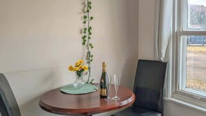 Un comedor con una mesa redonda de madera, dos sillas negras, una botella de champán y dos copas. Se ve una bombilla colgante y una planta de hiedra.
