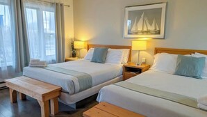 Habitación de hotel con dos camas, bancos de madera, un cuadro de veleros y una ventana con cortinas.