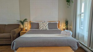 Ein ordentlich eingerichtetes Schlafzimmer mit einem großen Bett, Nachttischen mit Lampen, einer Bank und Blick ins Freie durch ein Fenster.