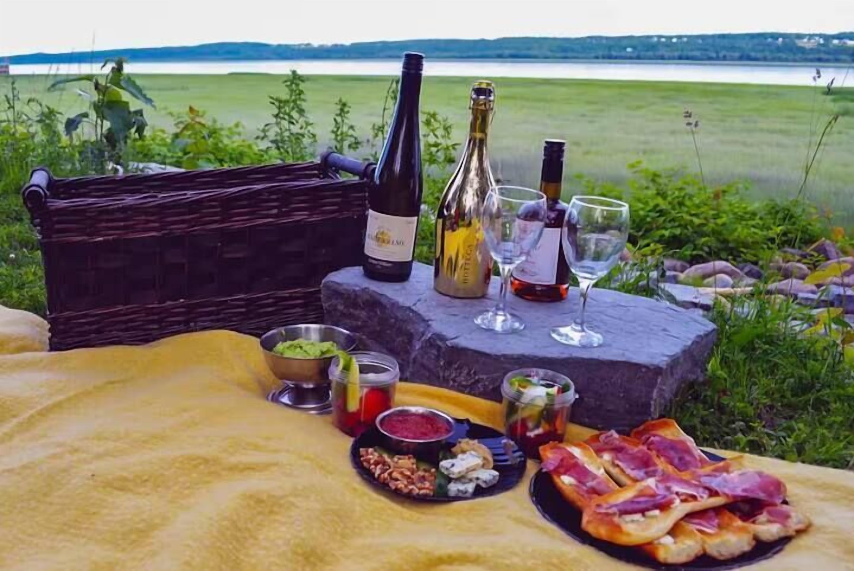 A picnic setup with a basket, bottles, glasses, and food on a blanket by a lake.