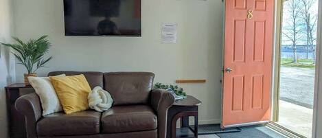 A living room with a brown leather sofa, a flat-screen TV, a wooden sign, and a door with a number.