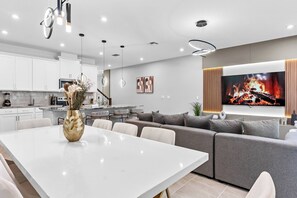 Una cocina moderna con armarios blancos, una zona de comedor con mesa blanca y un sofá gris.
