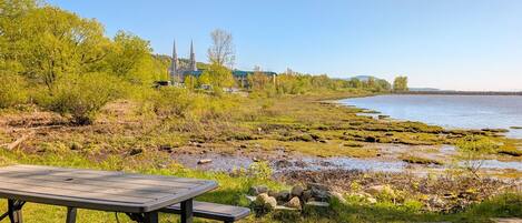 Picnicområde ved en flod med et træbord og bænke, omgivet af græsarealer og udsigt til en kirke og bakker i det fjerne.