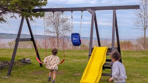 Spielplatz mit Schaukeln, einer gelben Rutsche und einem blauen Hängestuhl, umgeben von Wiese, Bäumen und einem See im Hintergrund.