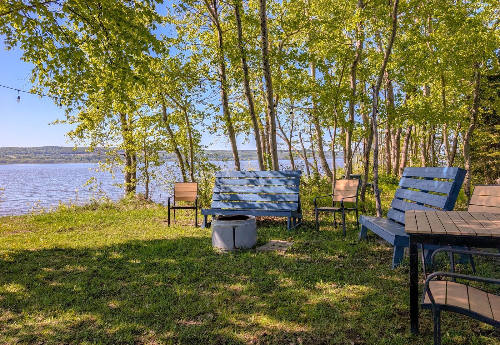An outdoor seating area with benches and chairs by a lake, surrounded by trees.
