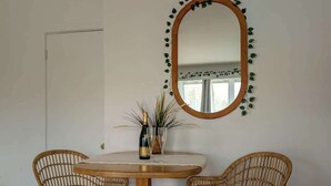 Una habitación con una mesa redonda, dos sillas de mimbre, un espejo con borde de hojas y una botella de champán sobre la mesa.