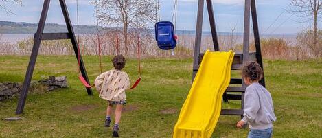 Spielplatz mit Schaukeln, einer gelben Rutsche und einem blauen Hängestuhl, umgeben von Wiese, Bäumen und einem See im Hintergrund.
