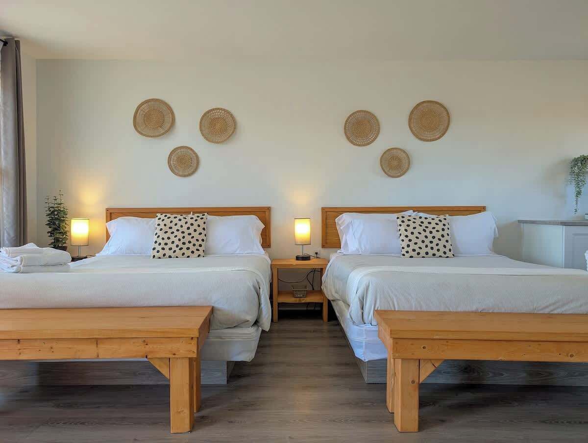 Habitación de hotel con dos camas, un banco de madera, una mesita de noche y adornos tejidos en la pared.