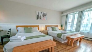 A hotel room with two wooden beds, a bench, a bedside table, and a window with curtains.