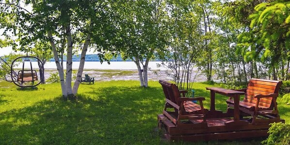 A wooden deck with a swing, chairs, and a table by a lake.