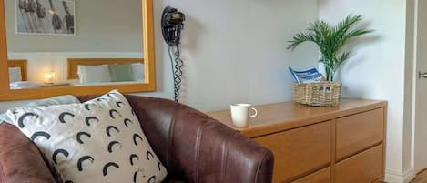 Olohuoneessa on ruskea nahkasohva, valkoinen koristetyyny, jossa on mustia puolikuun kuvioita, kirja, valkoinen muki, puinen kaappi, pieni ruukkukasvi ja seinälle asennettu televisio.