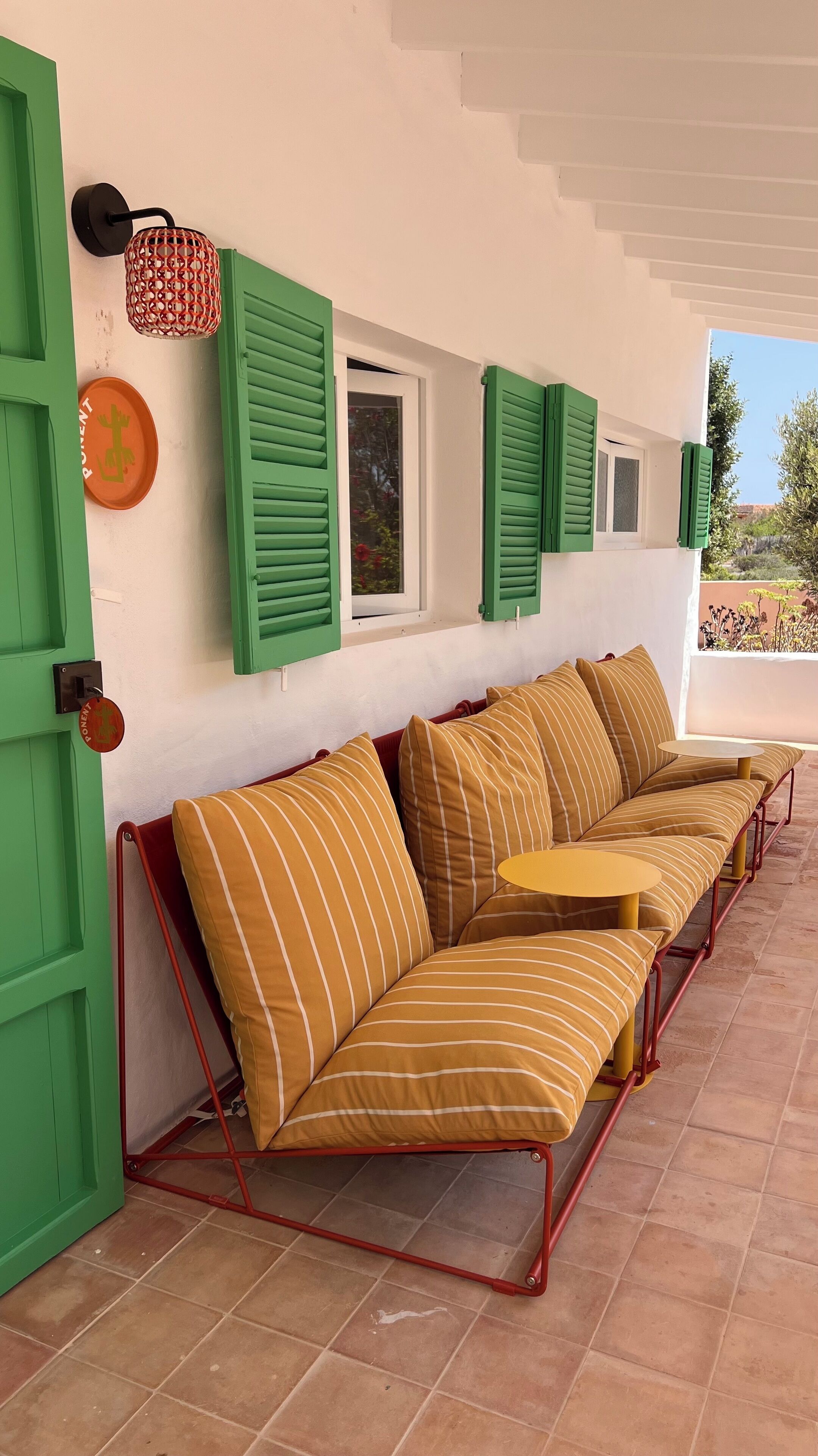 Un espacio de estar al aire libre cubierto con contraventanas verdes, un sofá de estructura metálica roja y una pequeña mesa.
