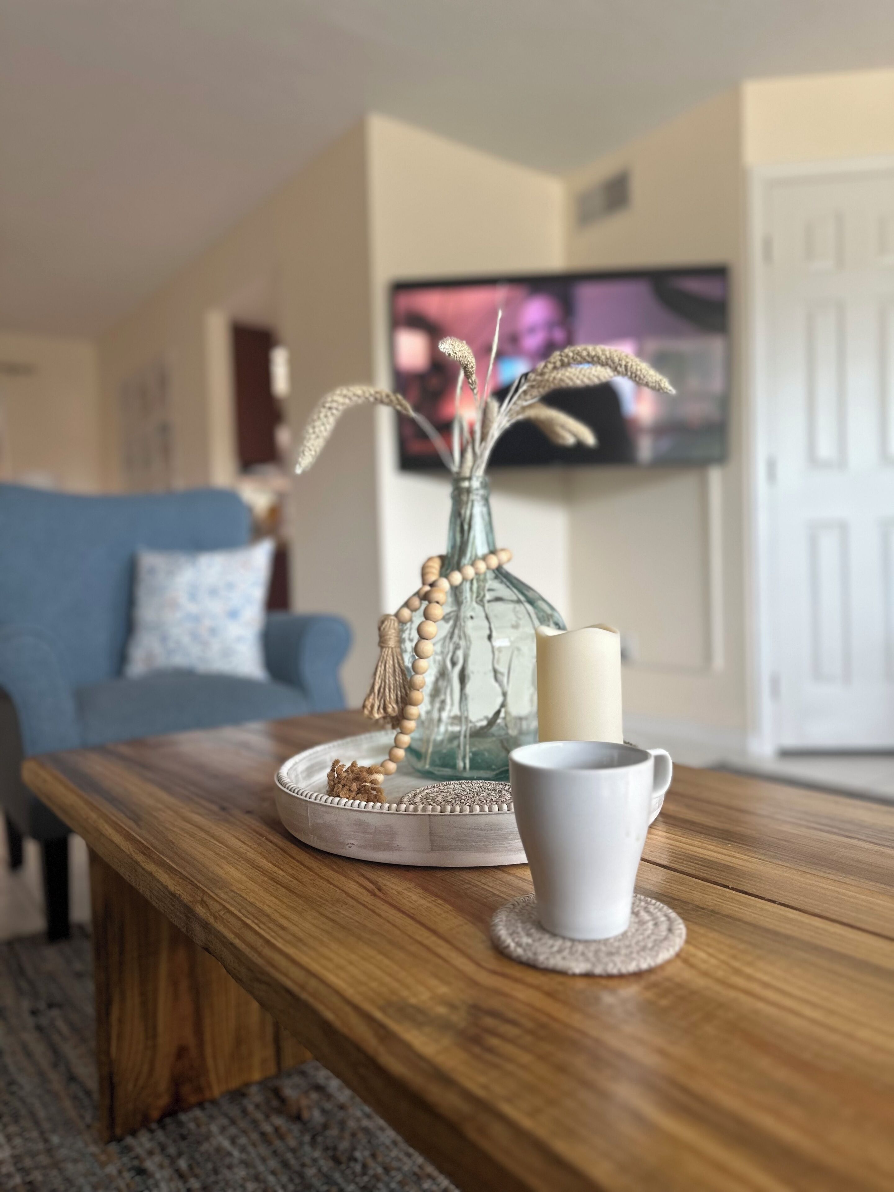 A wooden coffee table with a white mug, a glass vase with dried plants, and a decorative beaded necklace.
