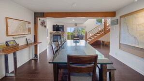 A spacious dining area with a long table and chairs, a fireplace, and a staircase leading to an upper level. - Sunset Point - Coos Bay, OR (Coos Bay)