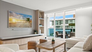 En moderne stue med et stort fjernsyn, der viser 'LUXURY RENTALS MIAMI BEACH', et træsofabord, hvide sofaer og udsigt til en bygning gennem store glasdøre.