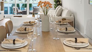 Et spiseområde med et langt bord dækket til seks personer, hvide tallerkener, klare vinglas og beige servietter. En vase med orange blomster står på bordet, og der er udsigt til en bygning med grønt udenfor vinduet.