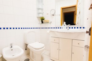 Ein Badezimmer mit Toilette, Bidet und Waschbecken. Über dem Waschbecken befindet sich ein Spiegel und ein Ablagebereich mit Pflegeprodukten.