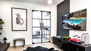 Een moderne woonkamer met een zwart leren vloerkleed, een lijst met abstract schilderij, een glazen deur die naar een keuken leidt en een grote tv aan de muur.