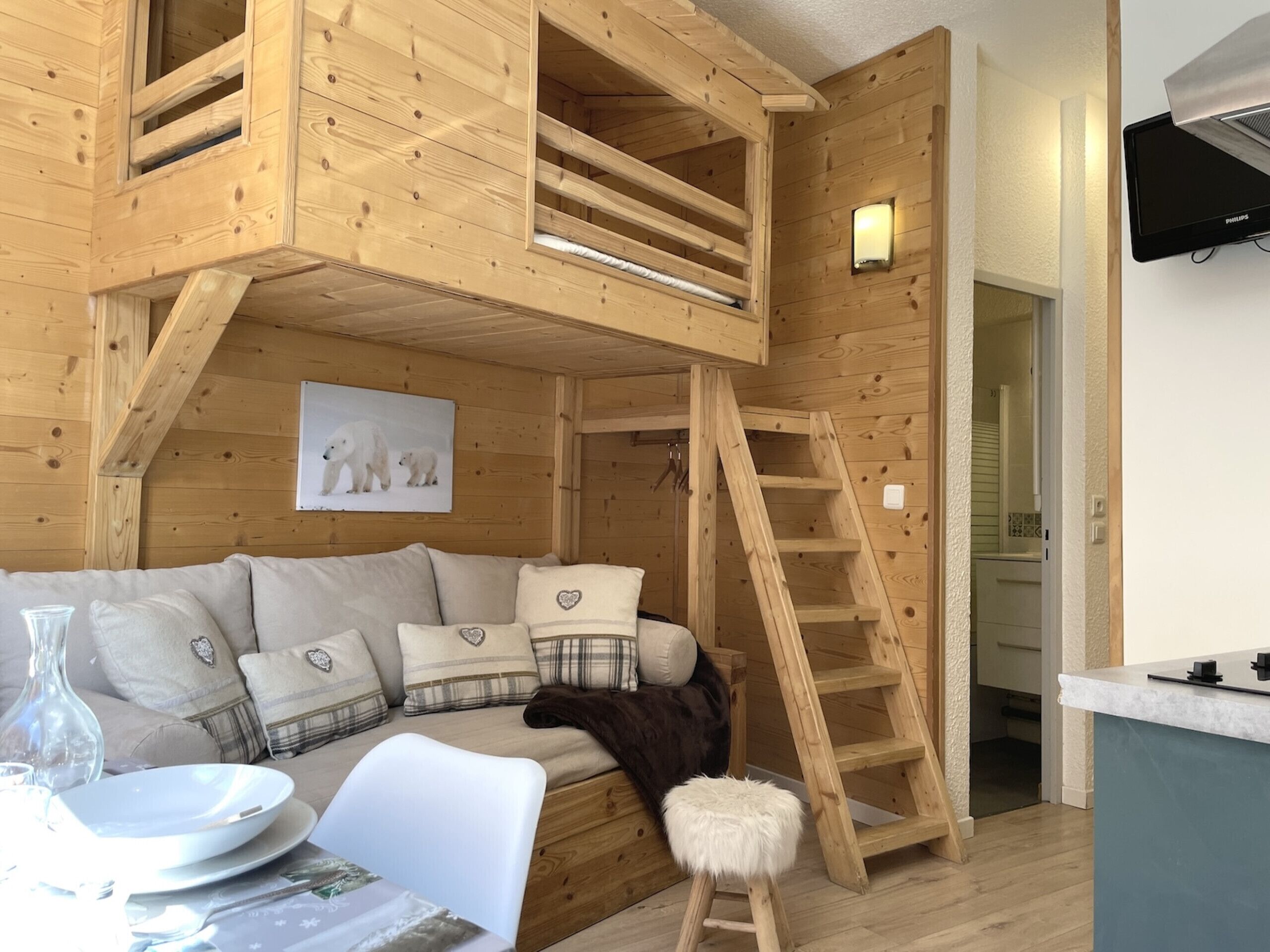 A cozy cabin interior with a loft bedroom, a wooden staircase, a sofa with heart-patterned cushions, and a painting of polar bears.