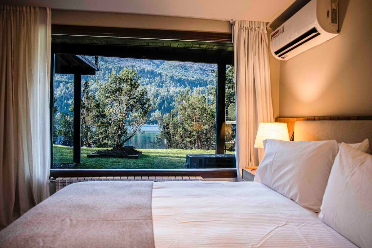 A hotel room with a large bed, a view of a lake and forest, and an air conditioning unit.