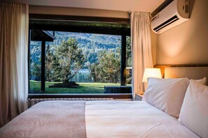 A hotel room with a large bed, a view of a lake and forest, and an air conditioning unit. - Auria Angostura (Villa La Angostura)