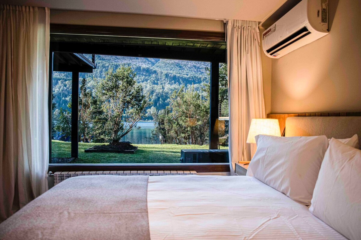 A hotel room with a large bed, a view of a lake and forest, and an air conditioning unit.