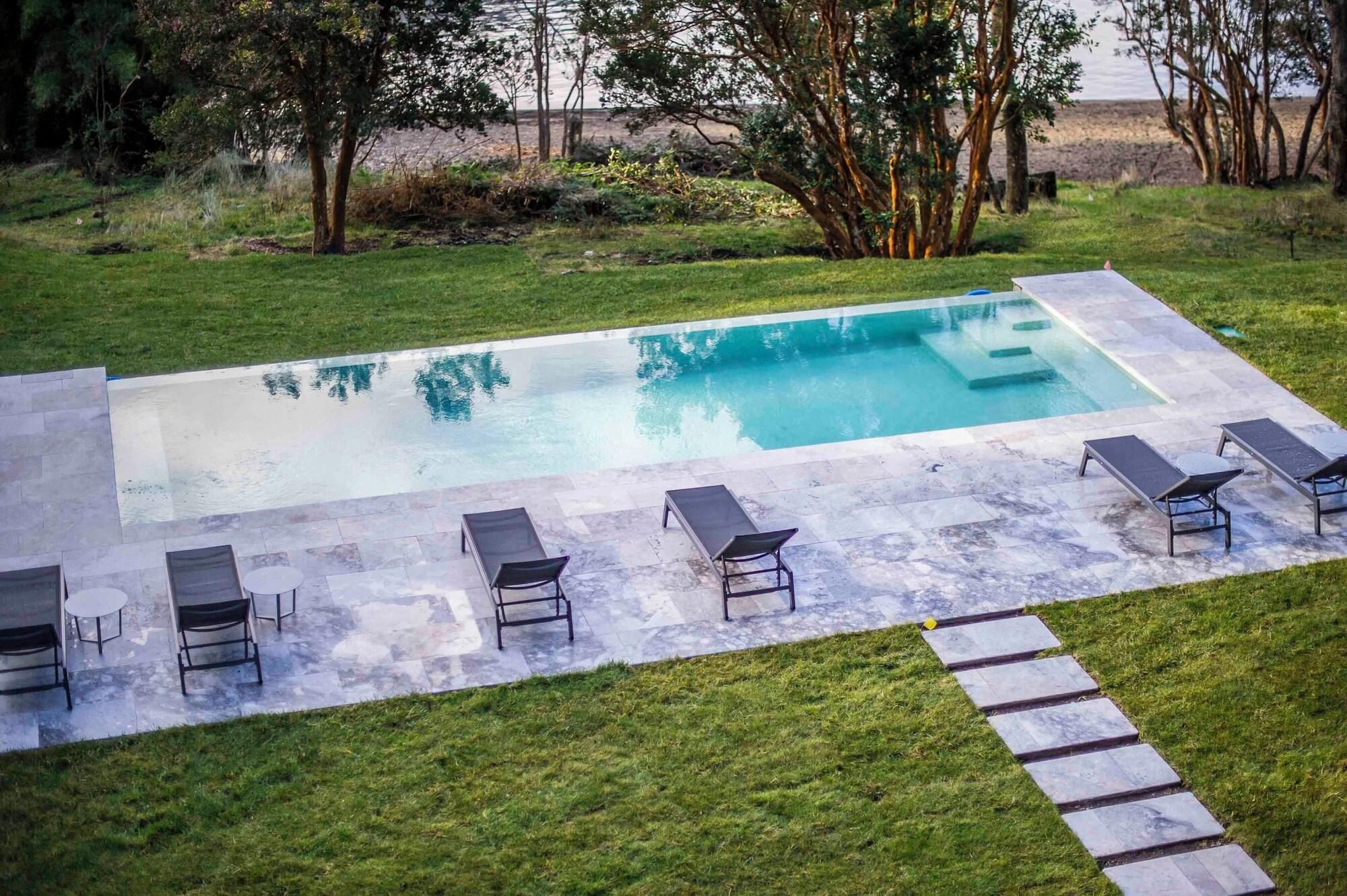 Una piscina rectangular con solárium empedrado, rodeada de césped y árboles.