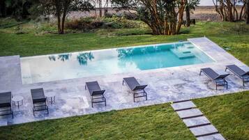 Una piscina rectangular con solárium empedrado, rodeada de césped y árboles.