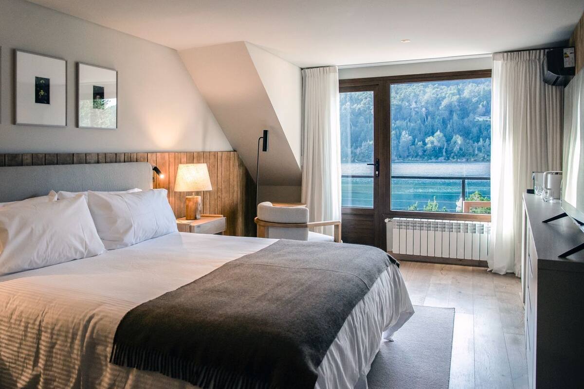 A hotel room with a large bed, a desk, a chair, a lamp, and a view of a lake.